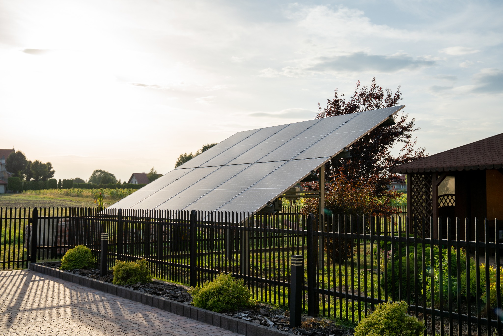 Panel fotowoltaiczny za czarnym ogrodzeniem, widok z boku. Małe krzewy dekoracyjne wzdłuż chodnika z kostki. W tle domy i drzewa.