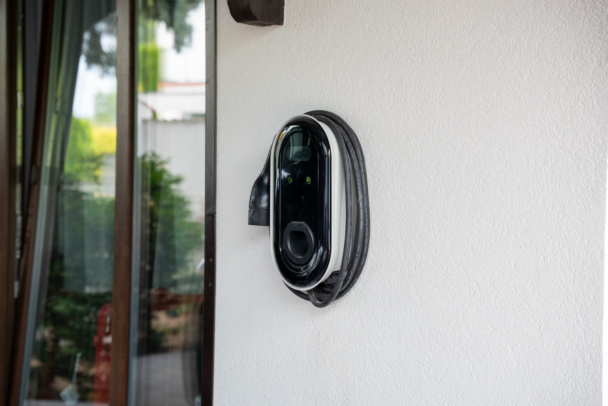 Naścienna stacja ładowania pojazdów elektrycznych z kablem, zamontowana na białej, teksturowanej ścianie zewnętrznej budynku, widoczna obok okna.