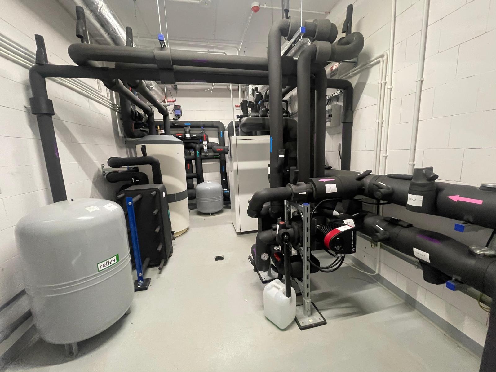 Węzeł cieplny w kotłowni: rury w izolacji, zbiorniki wyrównawcze Reflex, pompy obiegowe Grundfos, białe ściany z bloków, szara posadzka, instalacje pod sufitem.