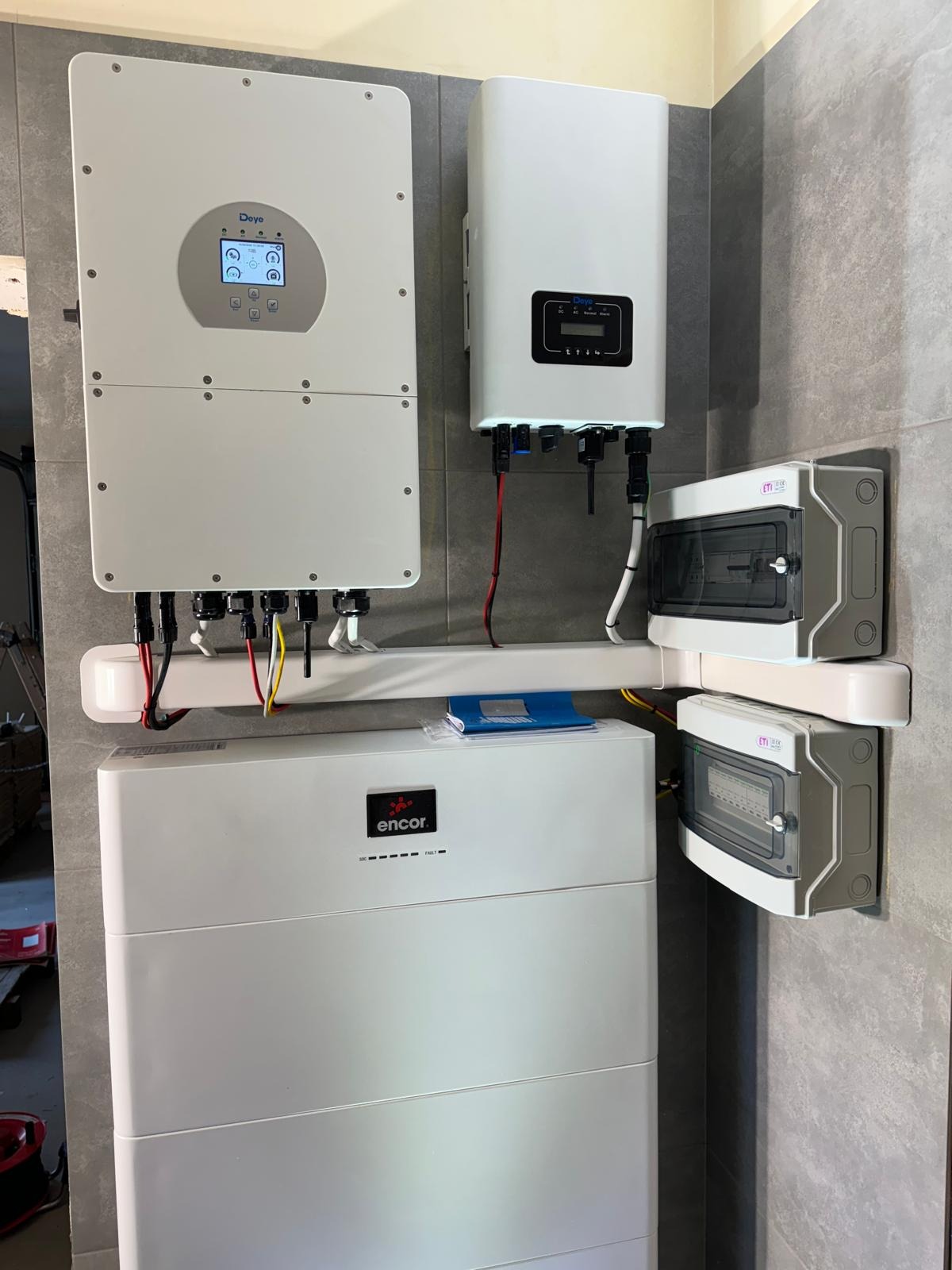 Zamontowany system magazynowania energii z falownikami na szarej ścianie. Widoczne przewody i zabezpieczenia elektryczne ETI. Kompletna instalacja w pomieszczeniu.