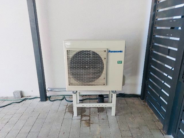 Biała jednostka zewnętrzna pompy ciepła Panasonic na metalowym stojaku, zainstalowana przy białej ścianie budynku i czarnym ogrodzeniu, widoczne przewody i kostka brukowa.