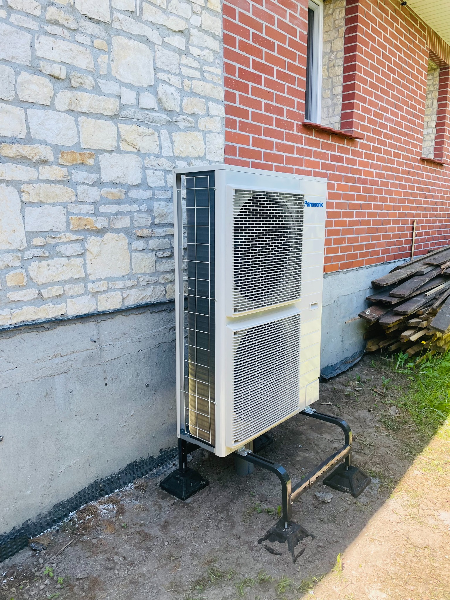 Zewnętrzna jednostka pompy ciepła Panasonic zamontowana na czarnej, metalowej podstawie przy ścianie budynku z cegły i kamienia, widoczne deski i trawa.