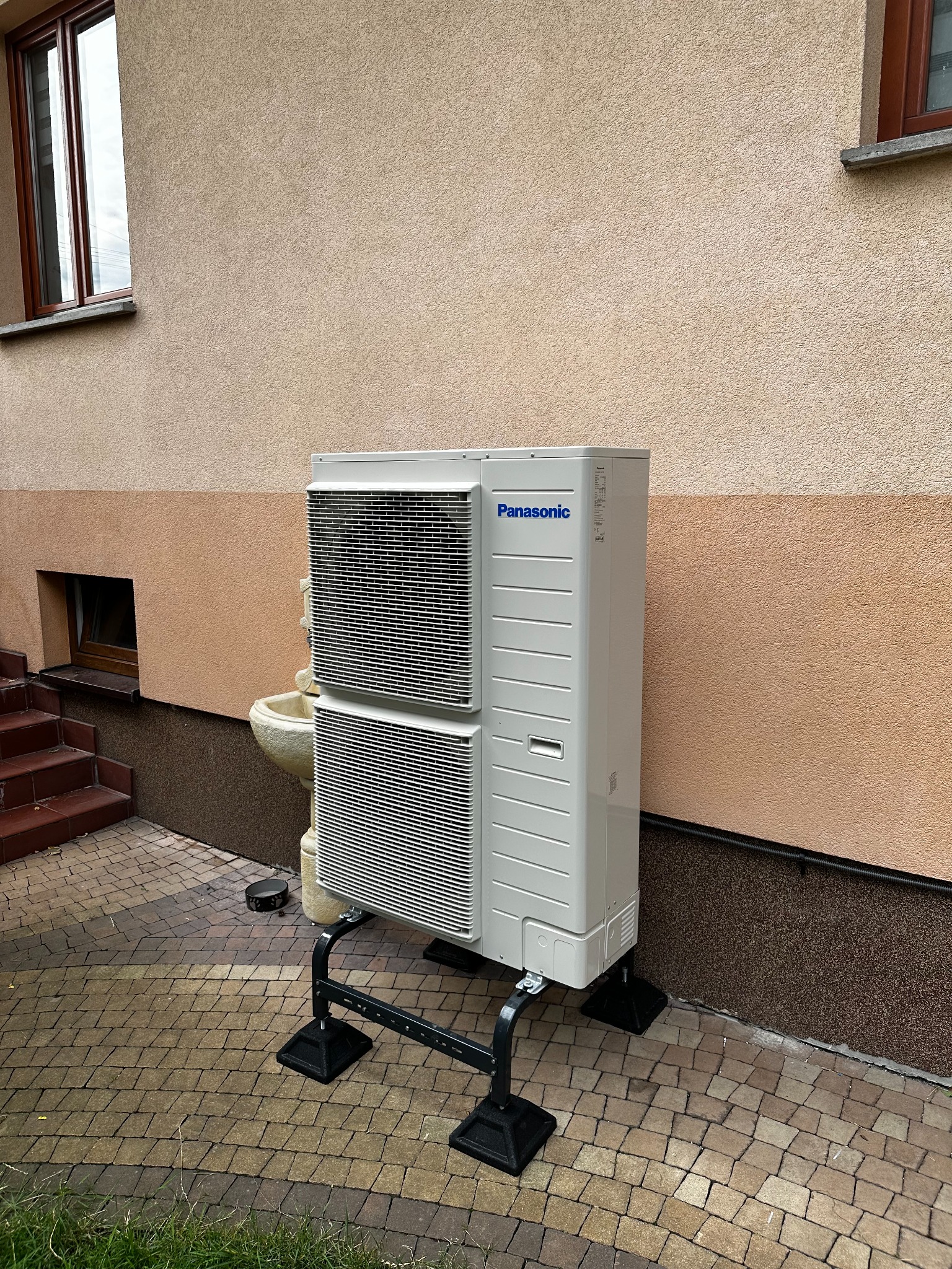 Biała jednostka zewnętrzna Panasonic na czarnym metalowym stelażu, ustawiona na kostce brukowej przy ścianie budynku z oknem i małą fontanną.
