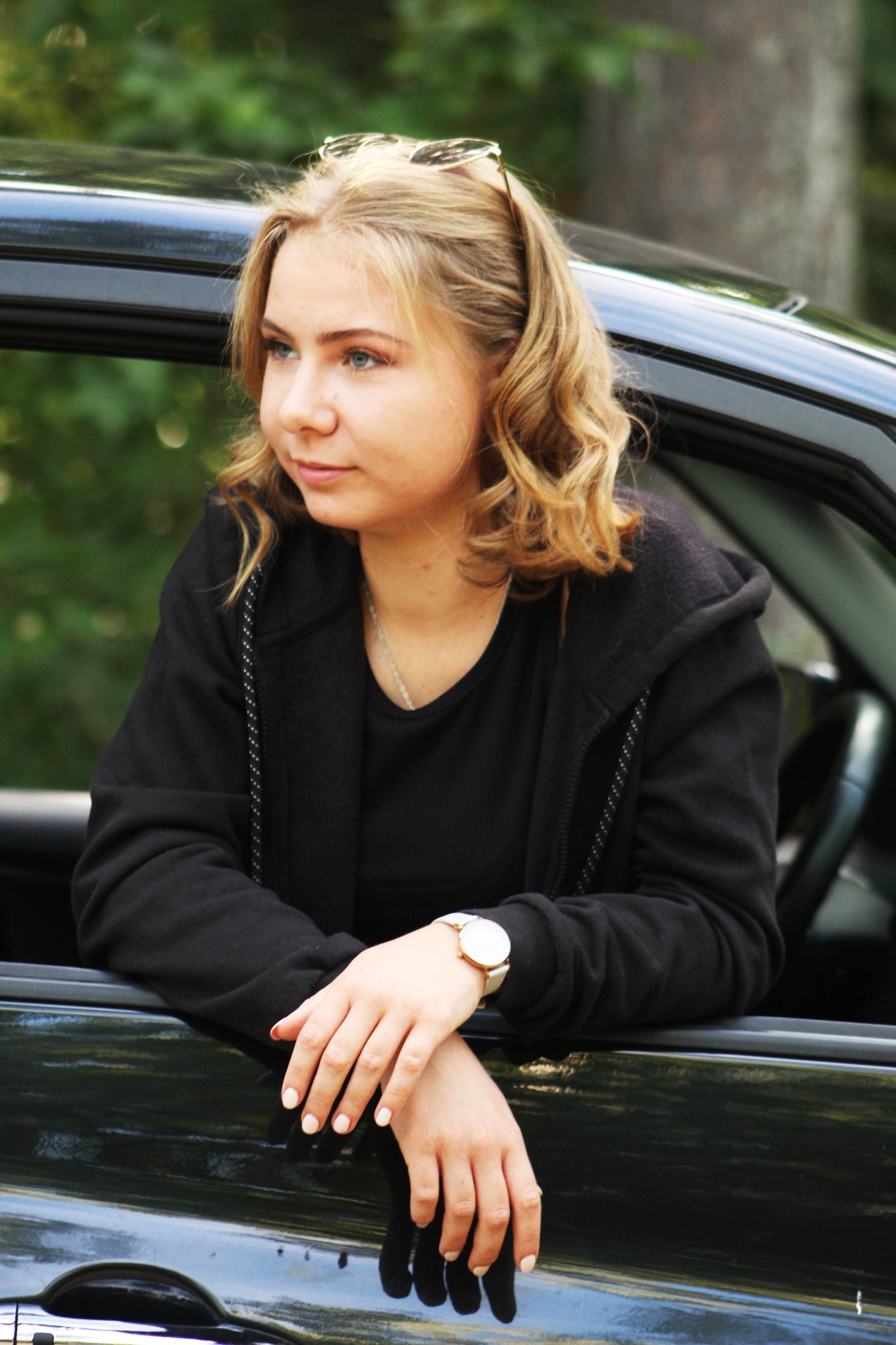 Portret młodej kobiety z blond włosami, opierającej się o czarny samochód, z okularami na głowie i zegarkiem na nadgarstku, spoglądającej w lewo.