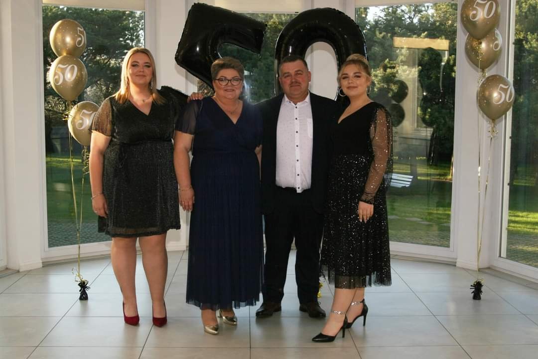 Rodzinne zdjęcie z okazji 50. urodzin, z balonami w kształcie cyfr i złotymi balonami z nadrukiem '50', w eleganckim wnętrzu z dużymi oknami.