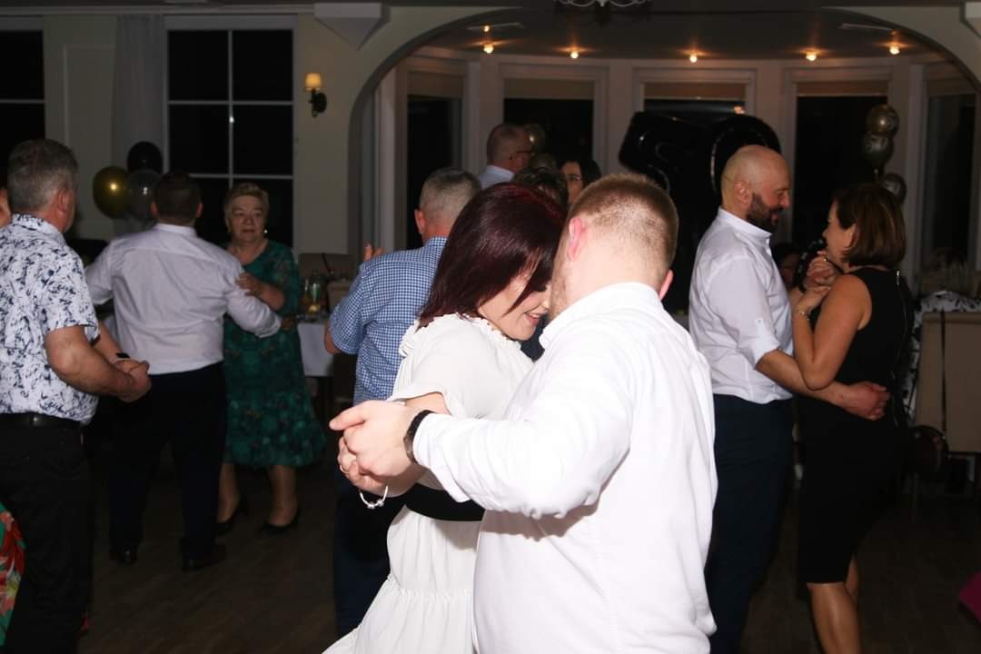 Para tańczy na parkiecie podczas imprezy, w tle widoczni inni goście i dekoracje balonowe.