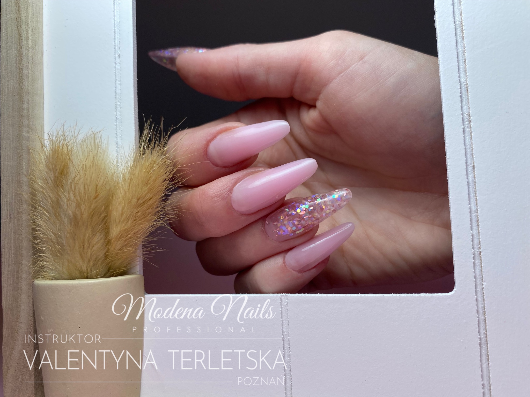 Dłoń z długimi, błyszczącymi paznokciami w odcieniach różu, prezentowana w białej ramce z logo studia Modena Nails, obok dekoracyjnej trawy w doniczce.