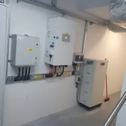 Instalacja magazynu energii: białe moduły bateryjne, skrzynki elektryczne i kable ułożone w korytkach na białej ścianie. Widoczna wentylacja pod sufitem i fragment schodów.