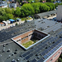 Gekon ENERGIA - Rozległa instalacja paneli słonecznych na dachu budynku z wewnętrznym dziedzińcem, widok z lotu ptaka, otoczenie miejskie z zielenią i budynkami w tle.