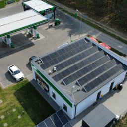 Gekon ENERGIA - Widok z góry na stację benzynową z panelami fotowoltaicznymi na dachu budynku. Obok dystrybutory paliw i zaparkowane samochody. Ujęcie z drona.