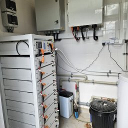 Gekon ENERGIA - Kompletna instalacja fotowoltaiczna w pomieszczeniu: bateria akumulatorów Deye, inwerter Gekon PV, rozdzielnice i białe płytki na ścianach. Widoczne rury instalacji.