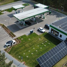 Gekon ENERGIA - Stacja paliw z panelami słonecznymi na dachu budynku i dodatkową instalacją na trawniku. Widok z lotu ptaka, słoneczny dzień. Samochody osobowe na parkingu.
