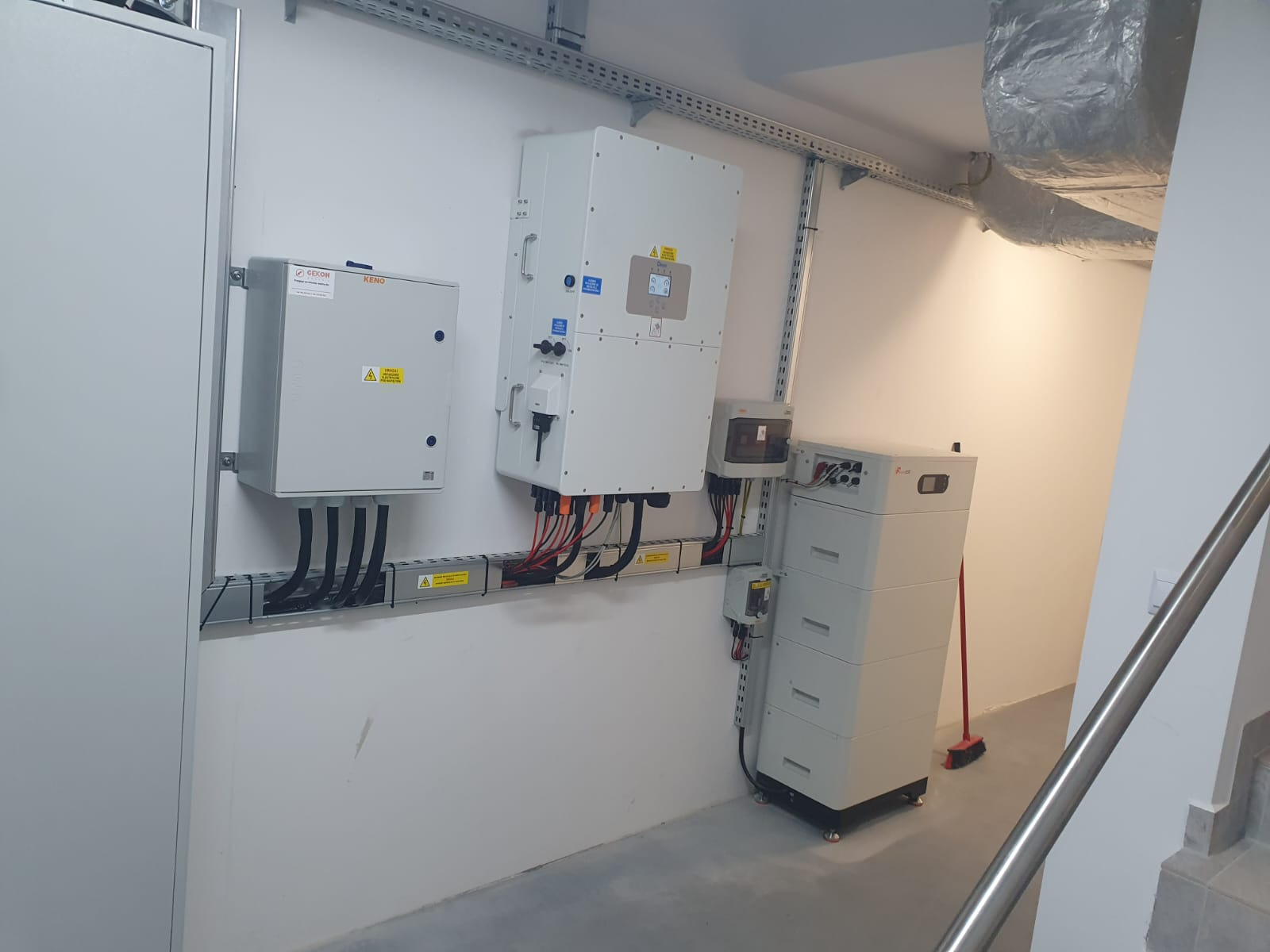 Instalacja magazynu energii: białe moduły bateryjne, skrzynki elektryczne i kable ułożone w korytkach na białej ścianie. Widoczna wentylacja pod sufitem i fragment schodów.