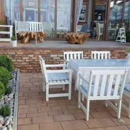 Zewnętrzna strefa relaksu z białymi meblami ogrodowymi, okrągłymi krzewami bukszpanu wzdłuż muru z cegły i dekoracyjnymi elementami z korzeni drzew na tarasie.