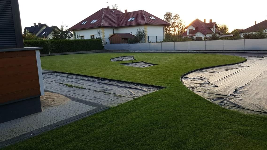 Zakładanie trawnika z rolki: widok na rozłożoną agrowłókninę i przygotowane podłoże pod trawnik, w tle dom jednorodzinny z czerwoną dachówką.