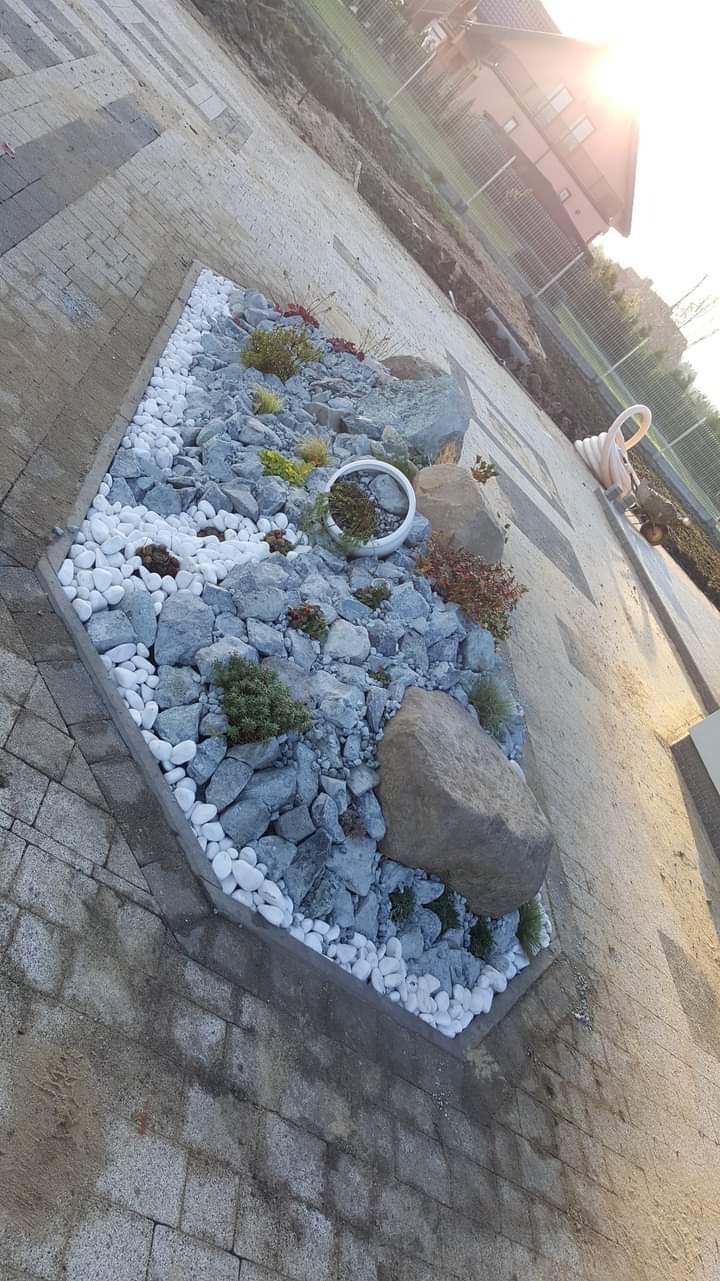 Kaskada kamieni w ogrodzie, białe otoczaki kontrastują z szarymi głazami, niskie rośliny ozdobne i donica ceramiczna, w tle kostka brukowa i dom jednorodzinny.