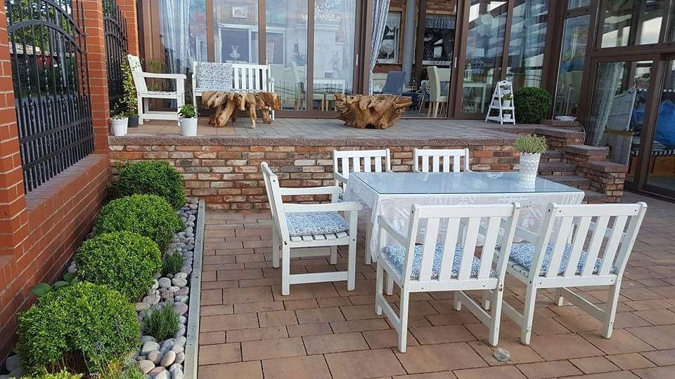 Zewnętrzna strefa relaksu z białymi meblami ogrodowymi, okrągłymi krzewami bukszpanu wzdłuż muru z cegły i dekoracyjnymi elementami z korzeni drzew na tarasie.