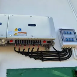 Biały falownik solarny Huawei zamontowany na ścianie, z podłączonymi czarnymi kablami i skrzynką bezpiecznikową po prawej stronie, na tle zielonej ściany.