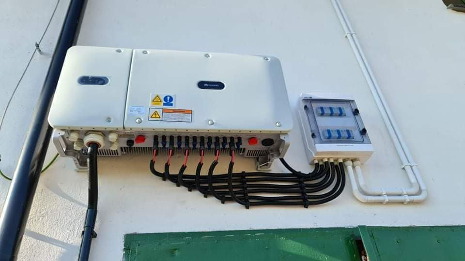 Biały falownik solarny Huawei zamontowany na ścianie, z podłączonymi czarnymi kablami i skrzynką bezpiecznikową po prawej stronie, na tle zielonej ściany.