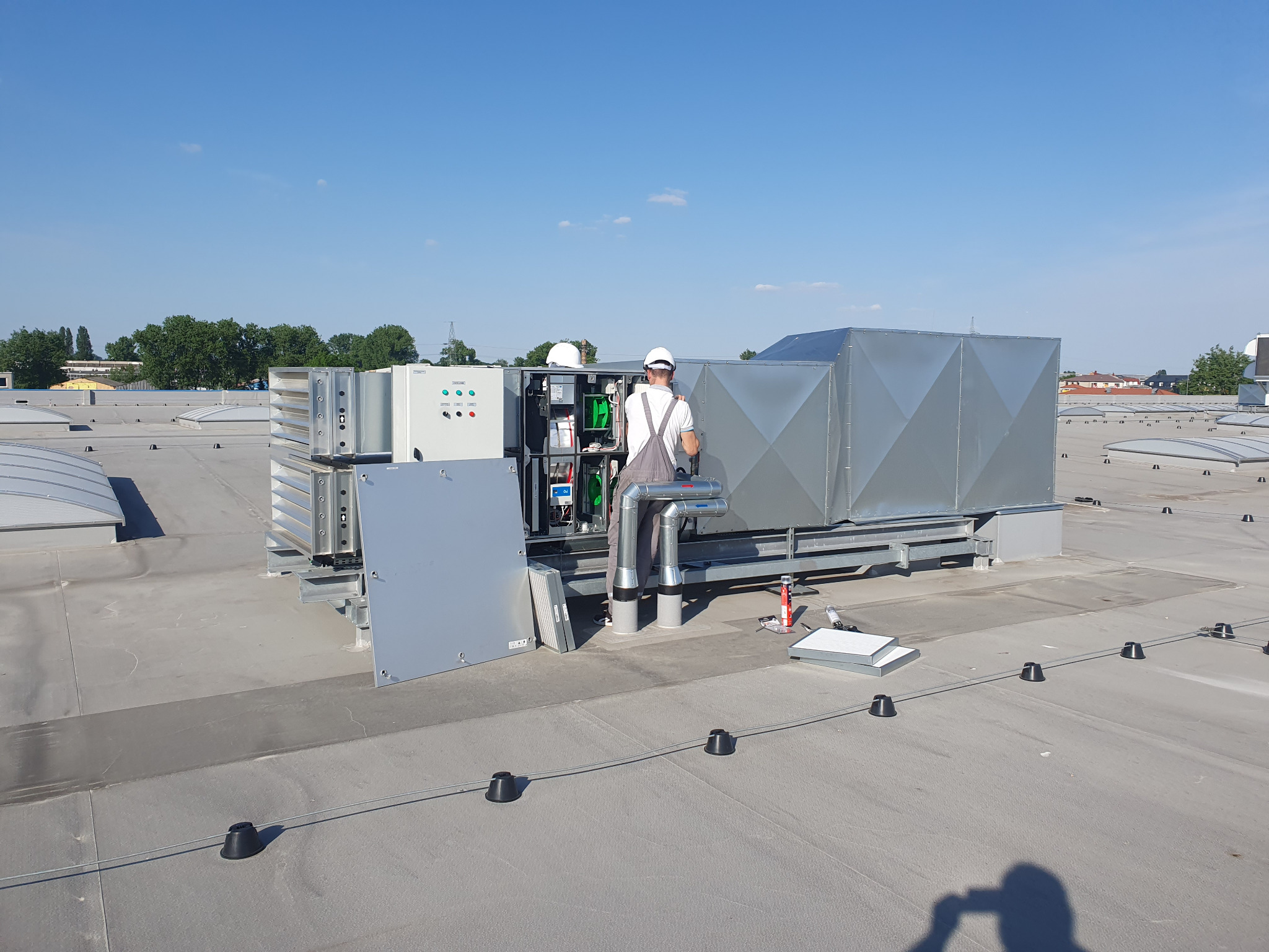 Montaż centrali wentylacyjnej z rekuperacją na dachu budynku, widoczni dwaj technicy w kaskach, instalacja w trakcie podłączania, elementy montażowe na dachu.