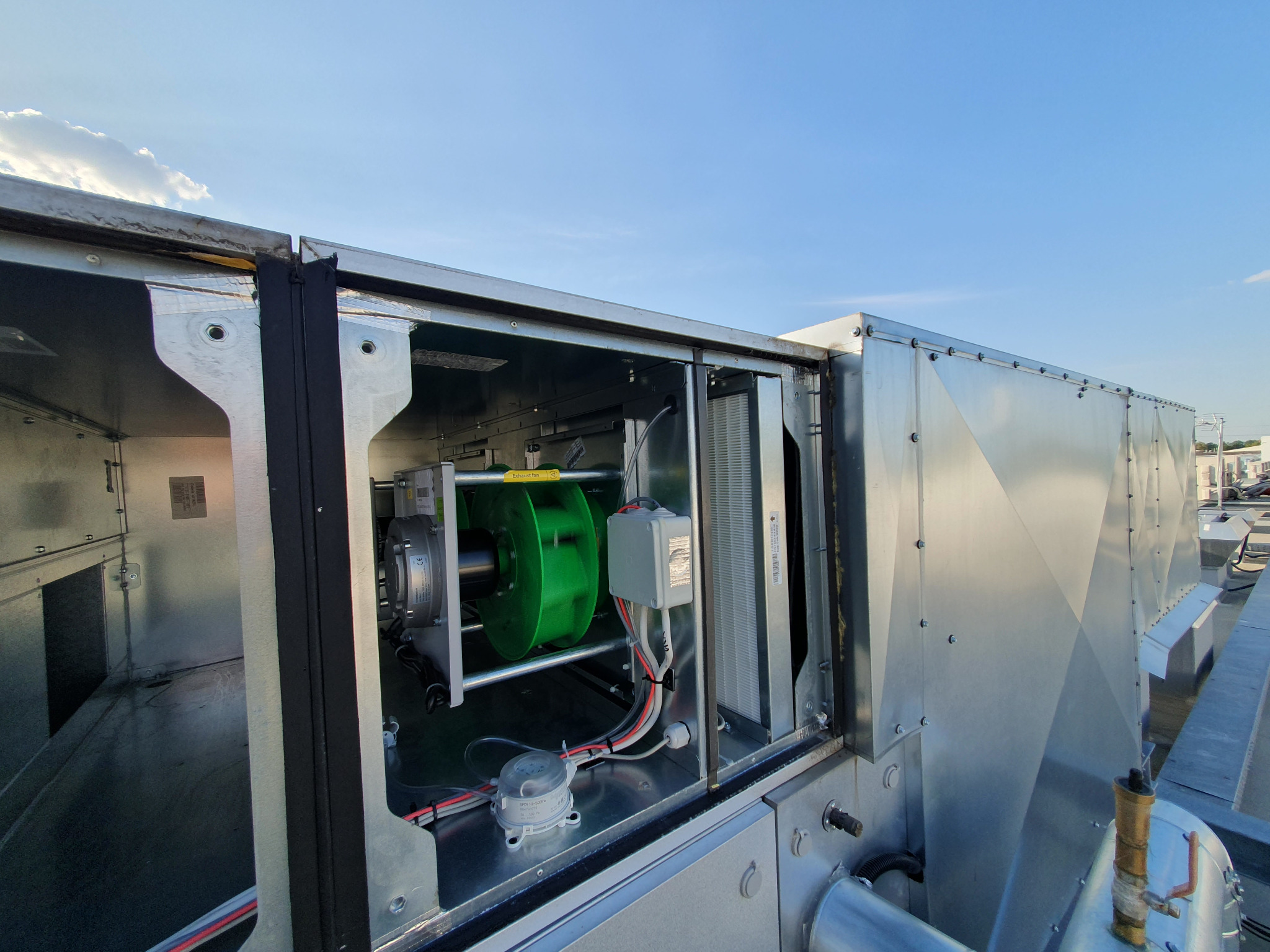 Centrala wentylacyjna z zielonym wentylatorem i filtrem, widoczna na dachu budynku w słoneczny dzień.