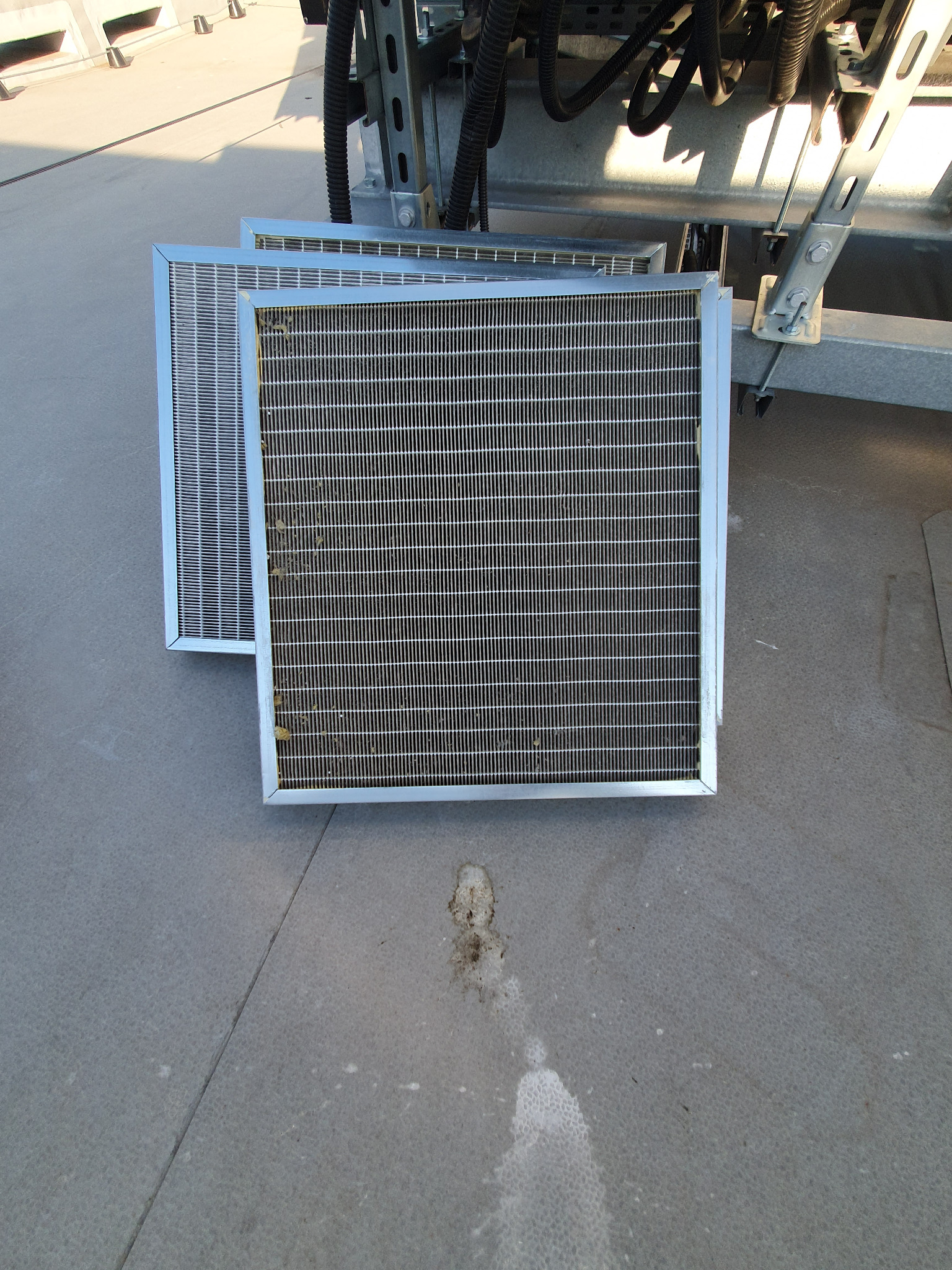 Dwa metalowe filtry kratkowe, jeden z widocznymi zabrudzeniami, leżące na szarej powierzchni, prawdopodobnie na dachu budynku, z fragmentem instalacji w tle.