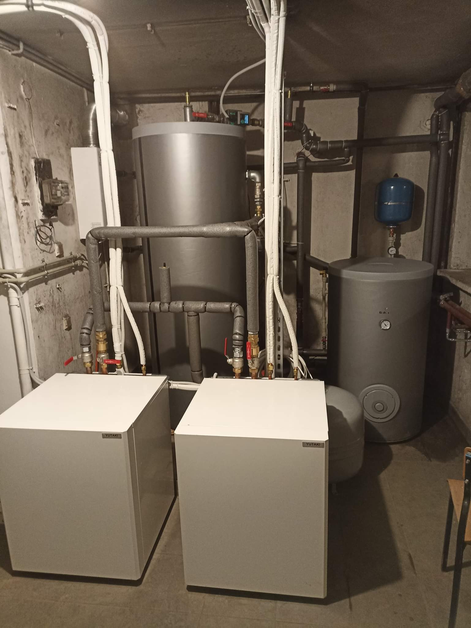Instalacja grzewcza w piwnicy: Dwa białe moduły YUTAKI, szary zbiornik akumulacyjny, rury w izolacji, manometry, zawory i niebieski zbiornik wyrównawczy.