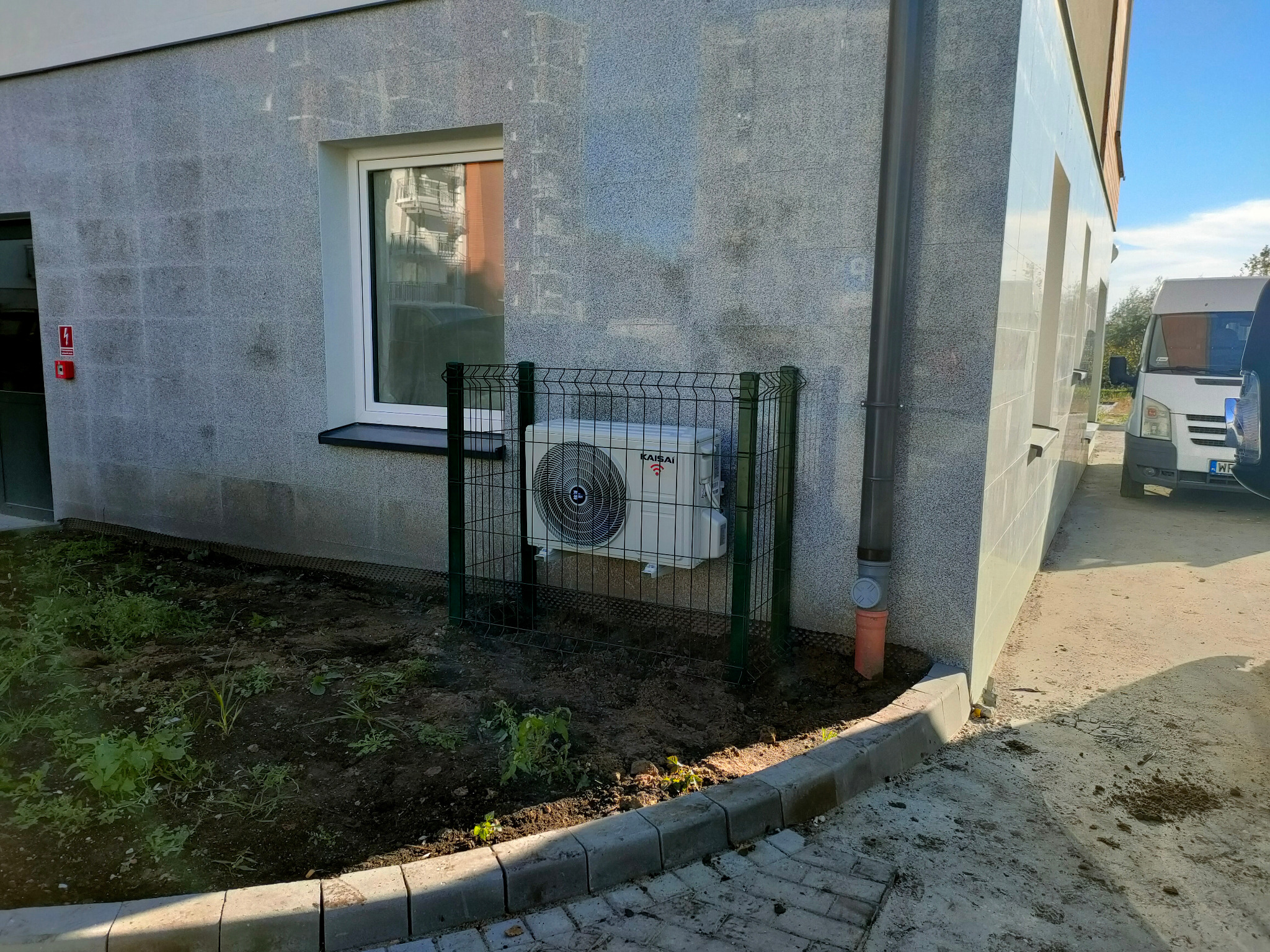 Zewnętrzna jednostka klimatyzatora Kaisai zamontowana przy ścianie budynku, zabezpieczona zielonym ogrodzeniem z siatki, z widocznymi elementami instalacji odprowadzającej skropliny.