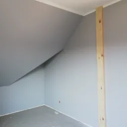 Pomalowane na szaro poddasze ze skośnym sufitem, białą listwą przysufitową i surową drewnianą belką przy ścianie.