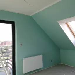 Pomalowane na turkusowo poddasze ze świeżo zamontowanym grzejnikiem, widoczne okno dachowe i fragment drabiny malarskiej przy oknie balkonowym.