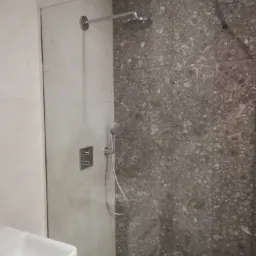 Nowoczesna kabina prysznicowa z szarymi płytkami imitującymi kamień, wyposażona w deszczownicę i panel sterujący, z widoczną umywalką w dolnym rogu.