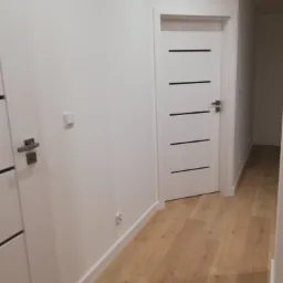 Jasne wnętrze mieszkania po remoncie: białe ściany, drewniana podłoga, białe drzwi z poziomymi czarnymi paskami.