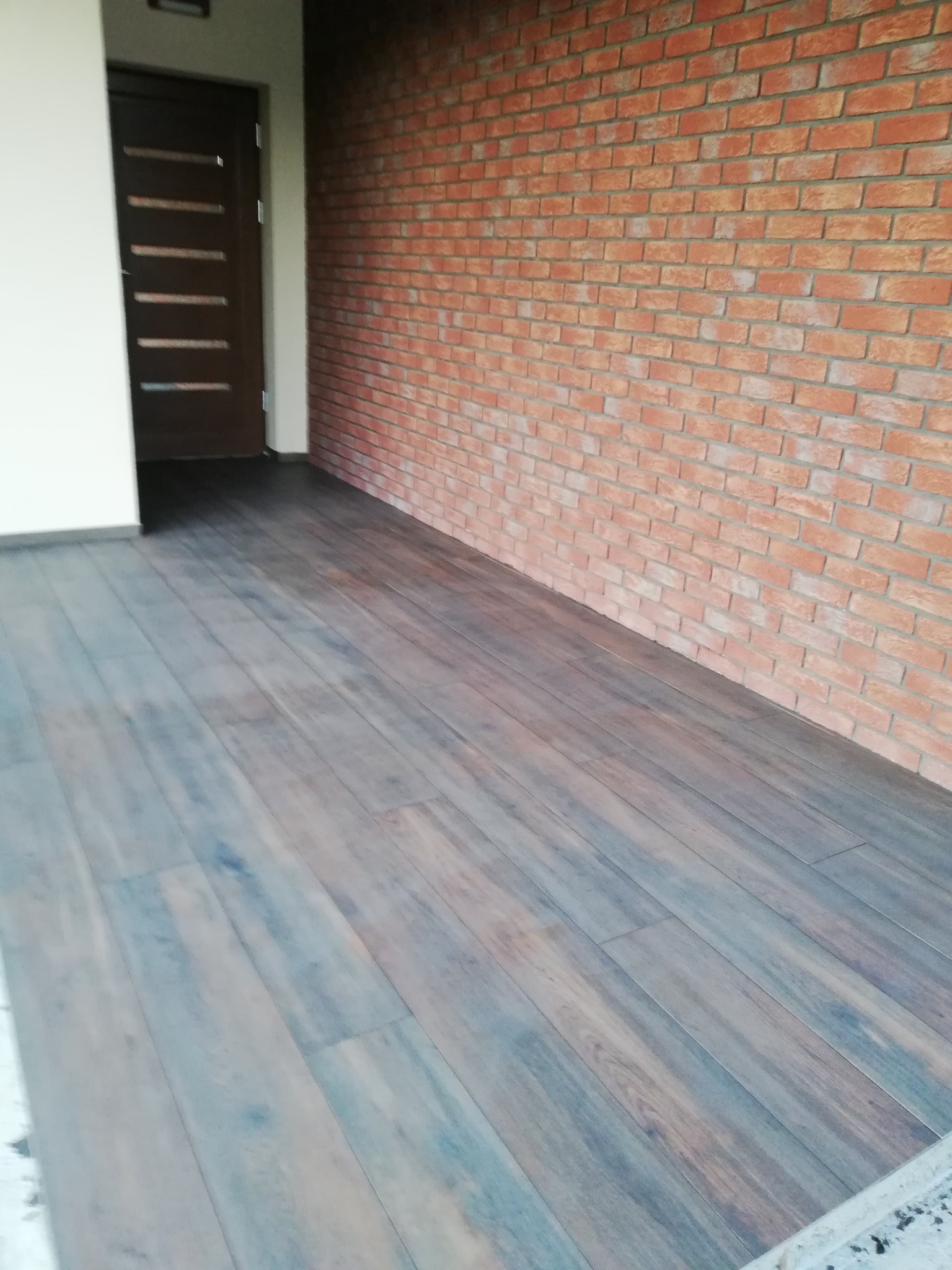 Ułożona podłoga z paneli imitujących drewno w odcieniach brązu i szarości, ściana wykończona cegłą klinkierową, widoczna stolarka drzwiowa w kolorze ciemnego brązu.