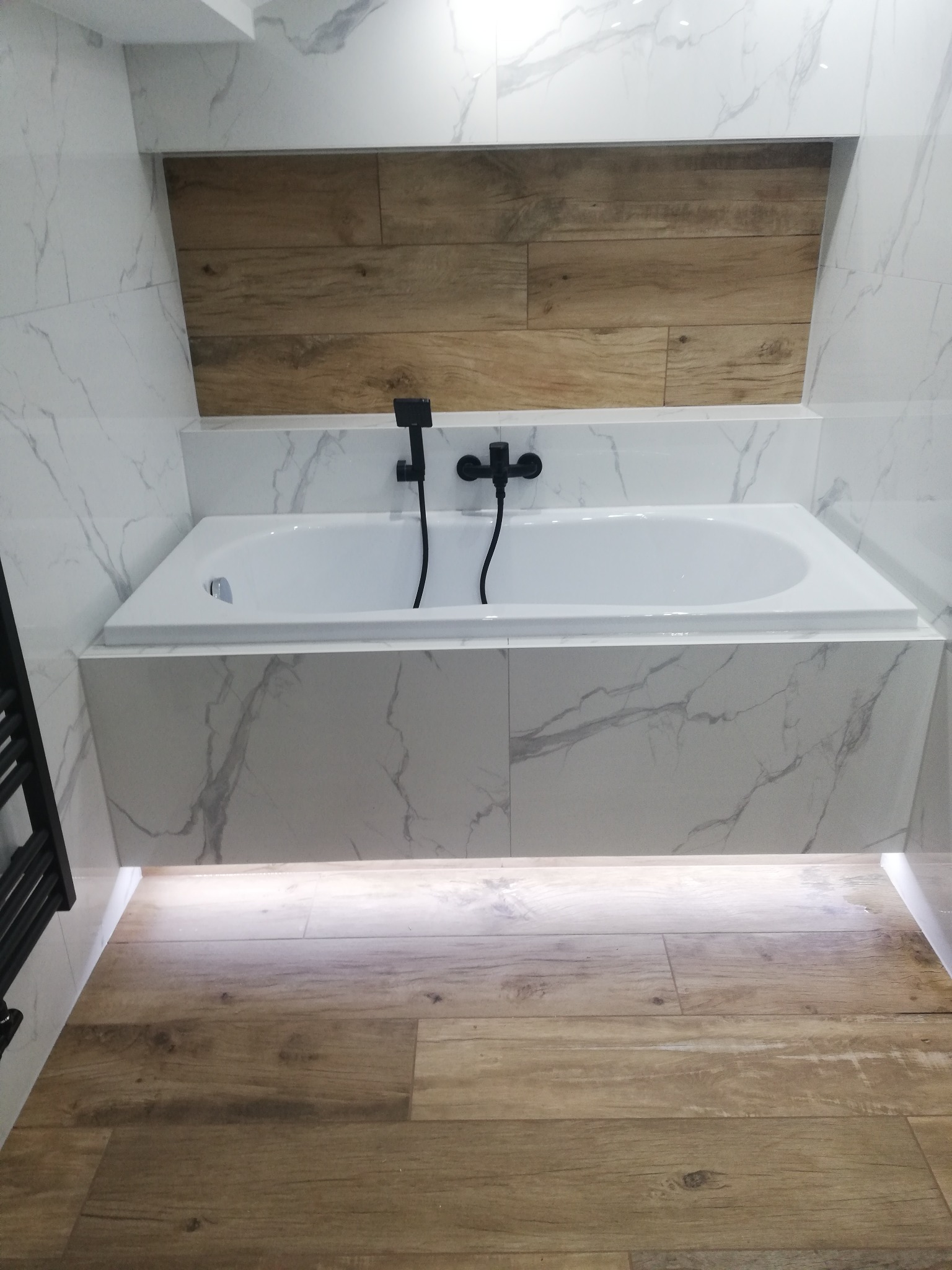 Nowoczesna łazienka z wanną obudowaną płytkami imitującymi marmur, czarna armatura i drewnopodobne płytki na ścianie wnęki oraz podświetlenie LED pod wanną.