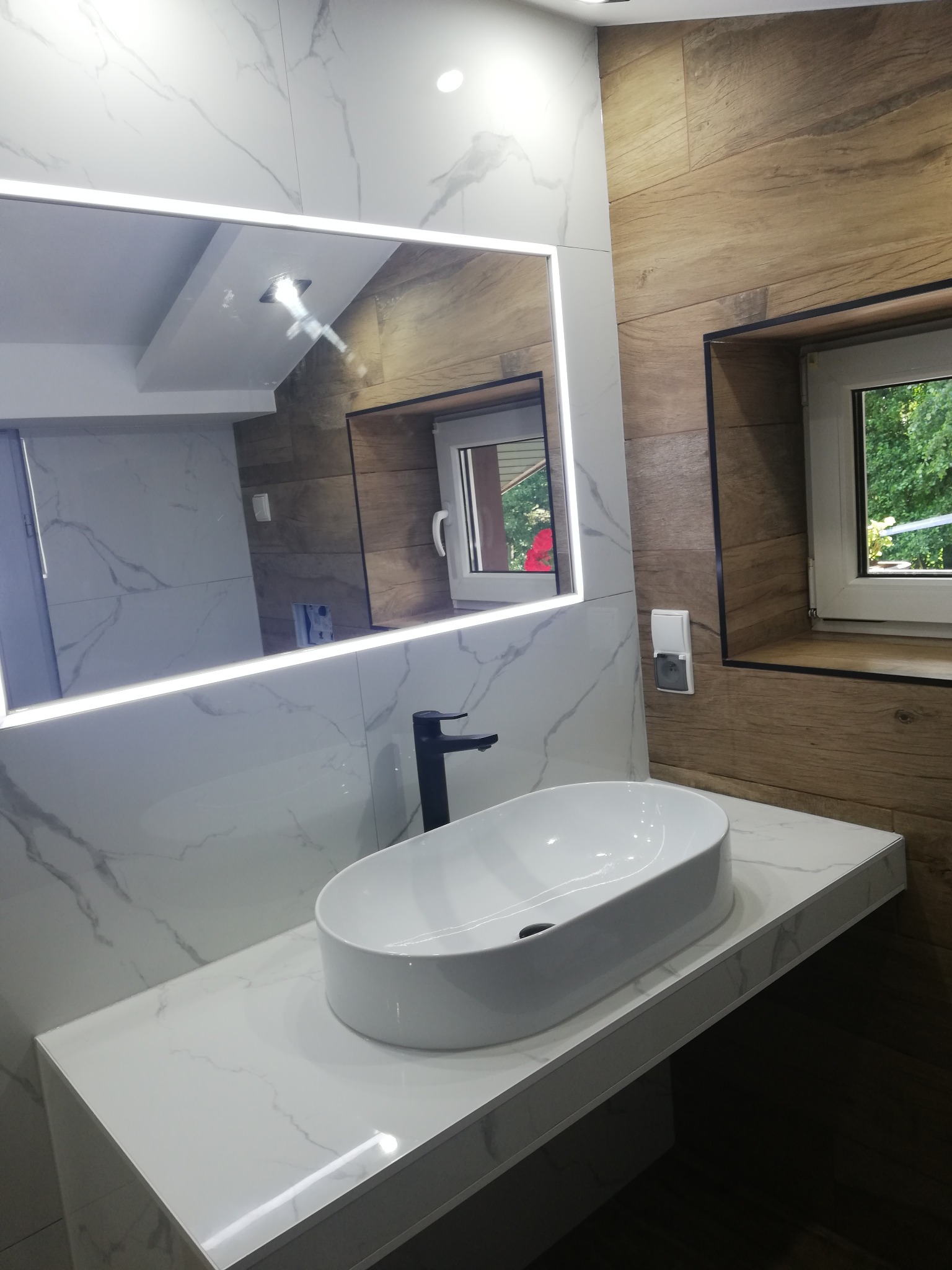 Nowoczesna łazienka z owalną umywalką nablatową, czarną armaturą i podświetlanym lustrem na tle marmurowej ściany i drewnopodobnych paneli ściennych z oknem.