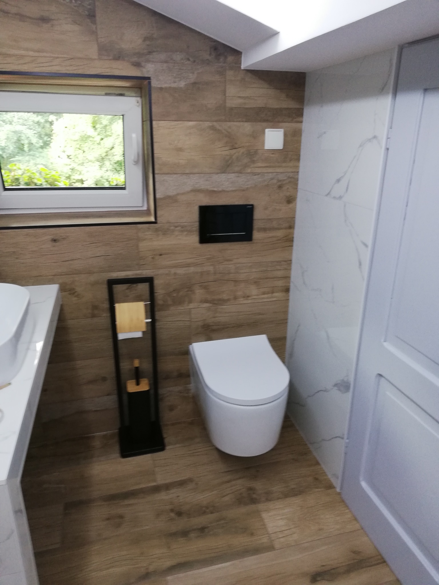 Nowoczesna łazienka z drewnopodobnymi płytkami na ścianach i podłodze, białą armaturą i oknem z widokiem na zieleń.