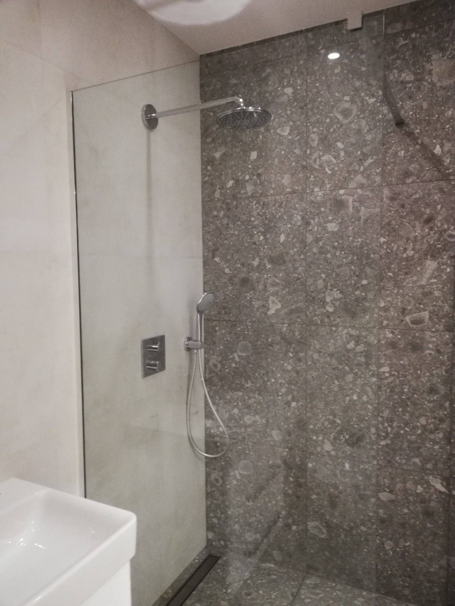 Nowoczesna kabina prysznicowa z szarymi płytkami imitującymi kamień, wyposażona w deszczownicę i panel sterujący, z widoczną umywalką w dolnym rogu.