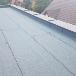 Widok z góry na płaski dach pokryty papą termozgrzewalną w kolorze szaro-niebieskim, z widocznymi łączeniami i obróbkami blacharskimi wzdłuż krawędzi, z domem jednorodzinnym w tle.