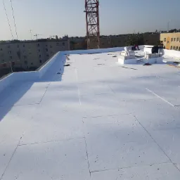 Białe płyty izolacyjne ułożone na płaskim dachu budynku wielorodzinnego, widoczni pracownicy w trakcie pracy, w tle dźwig i zabudowa miejska.