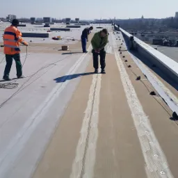 Prace na dachu płaskim: trzech dekarzy mocuje izolację, widoczne narzędzia i materiały izolacyjne, w tle panorama miasta.