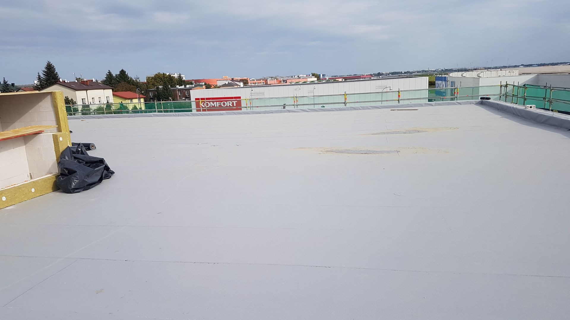 Płaski dach pokryty szarą membraną, widoczna izolacja przy krawędzi, w tle budynki miejskie i niebo.