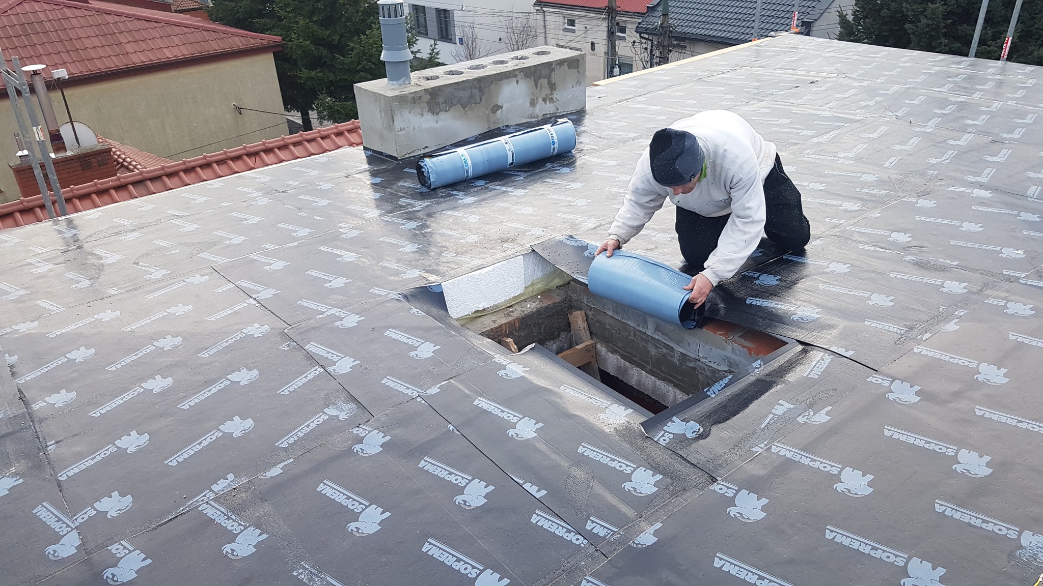Pracownik w trakcie układania hydroizolacji marki Soprema na dachu, widoczny otwór w dachu z drewnianą konstrukcją, w tle budynki mieszkalne i rolka materiału izolacyjnego.