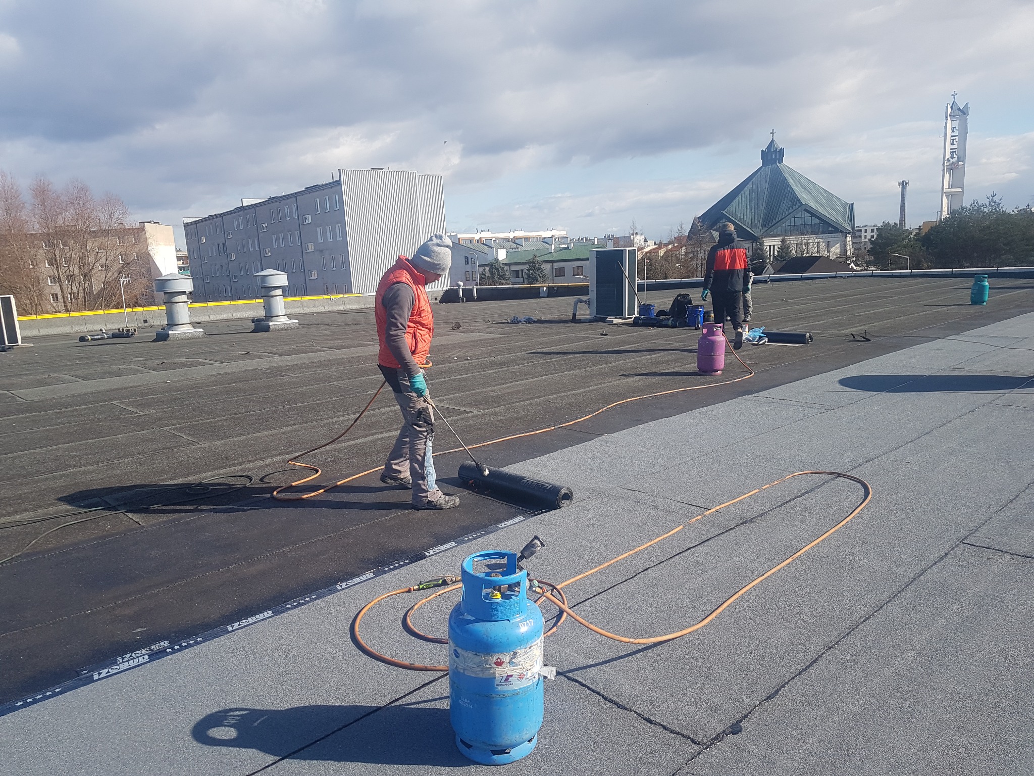 Praca na dachu: dekarz z palnikiem termicznym łączy papę bitumiczną na płaskim dachu, w tle zabudowa miejska i kościół.