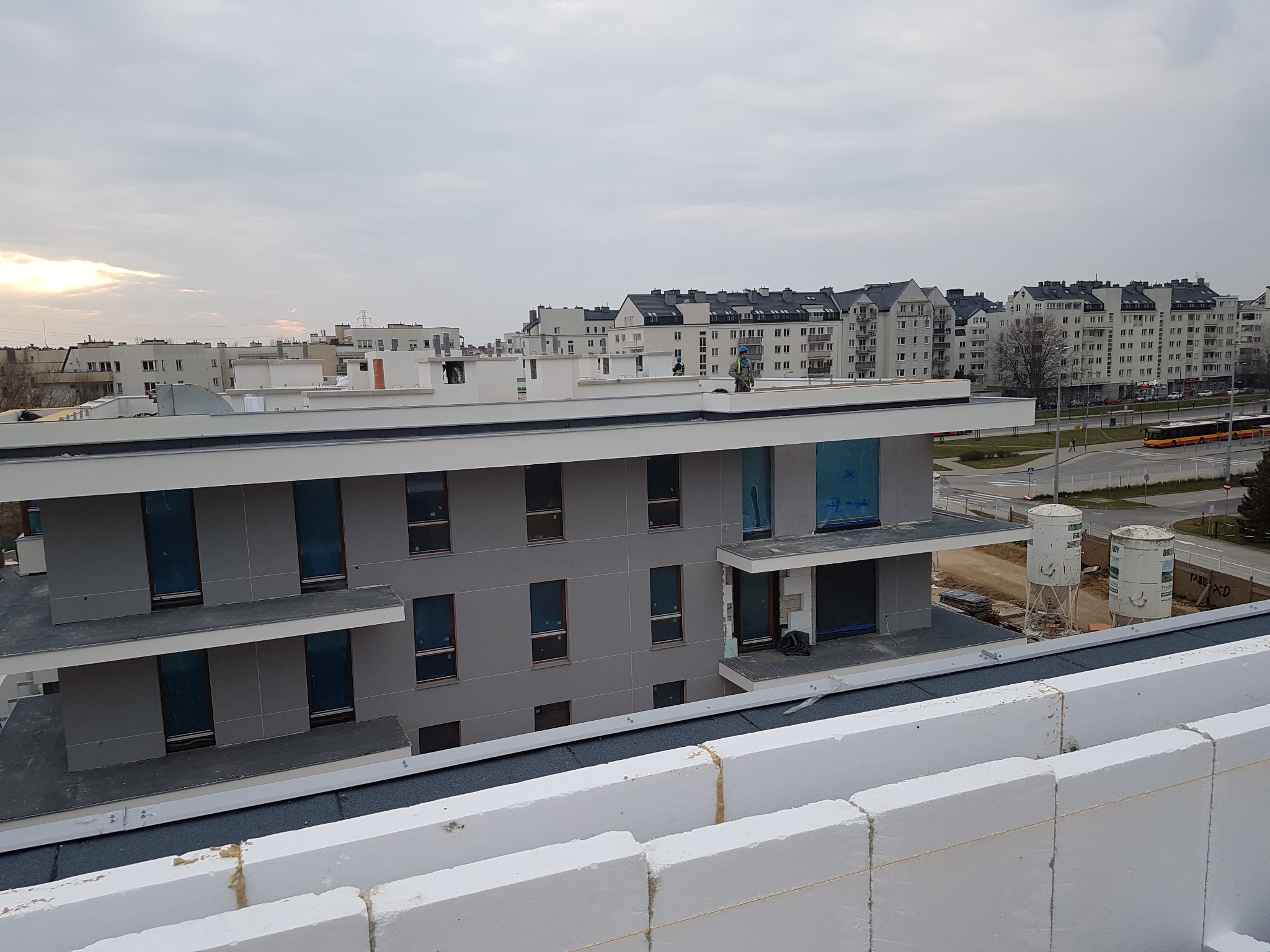 Nowoczesny budynek wielorodzinny z płaskim dachem w trakcie budowy, widoczne białe bloczki konstrukcyjne na pierwszym planie i robotnicy na dachu w oddali, szary budynek w tle z zabezpieczonymi...