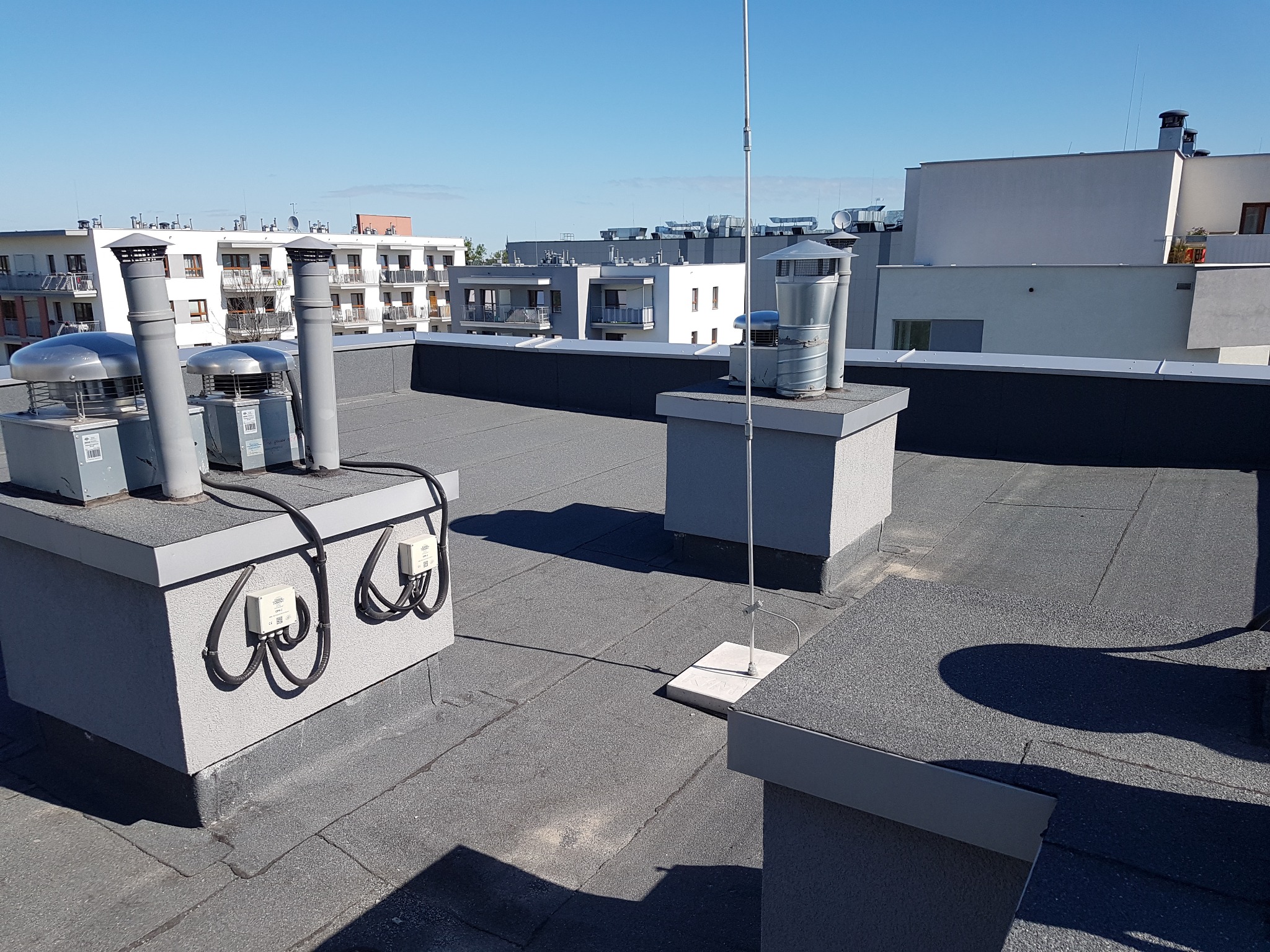 Płaski dach pokryty papą termozgrzewalną z widocznymi kominami wentylacyjnymi, instalacją odgromową oraz osprzętem elektrycznym, na tle zabudowy wielorodzinnej w słoneczny dzień.
