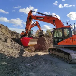 Dwa pomarańczowe, gąsienicowe, ciężkie Doosany pracują na placu budowy pod błękitnym niebem z chmurami, jeden z czerwoną łyżką koparki. W tle widać skarpę ziemi.