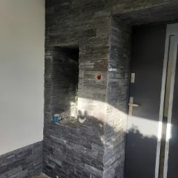 Wykończenie elewacji wejściowej kamieniem dekoracyjnym w odcieniach szarości, z wnęką na ozdoby i widoczną puszką elektryczną, sąsiadujące z nowoczesnymi drzwiami wejściowymi.