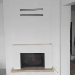 Nowoczesny, biały kominek z prostokątnymi wnękami wentylacyjnymi nad paleniskiem, widoczne ślady prac wykończeniowych na podłodze.