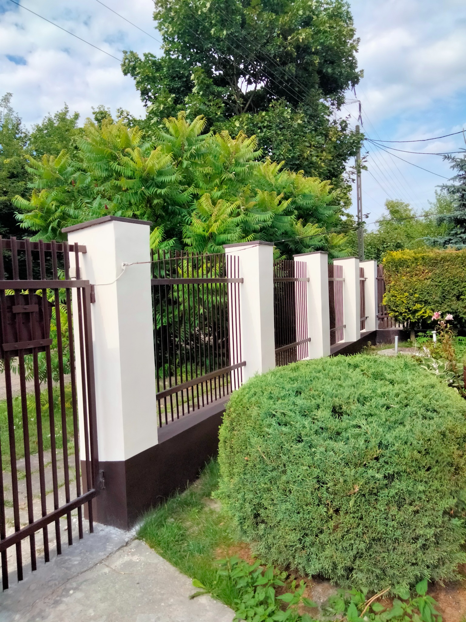 Nowoczesne ogrodzenie z metalowymi przęsłami i słupkami w kolorze beżowym i brązowym, otoczone zielenią ogrodu