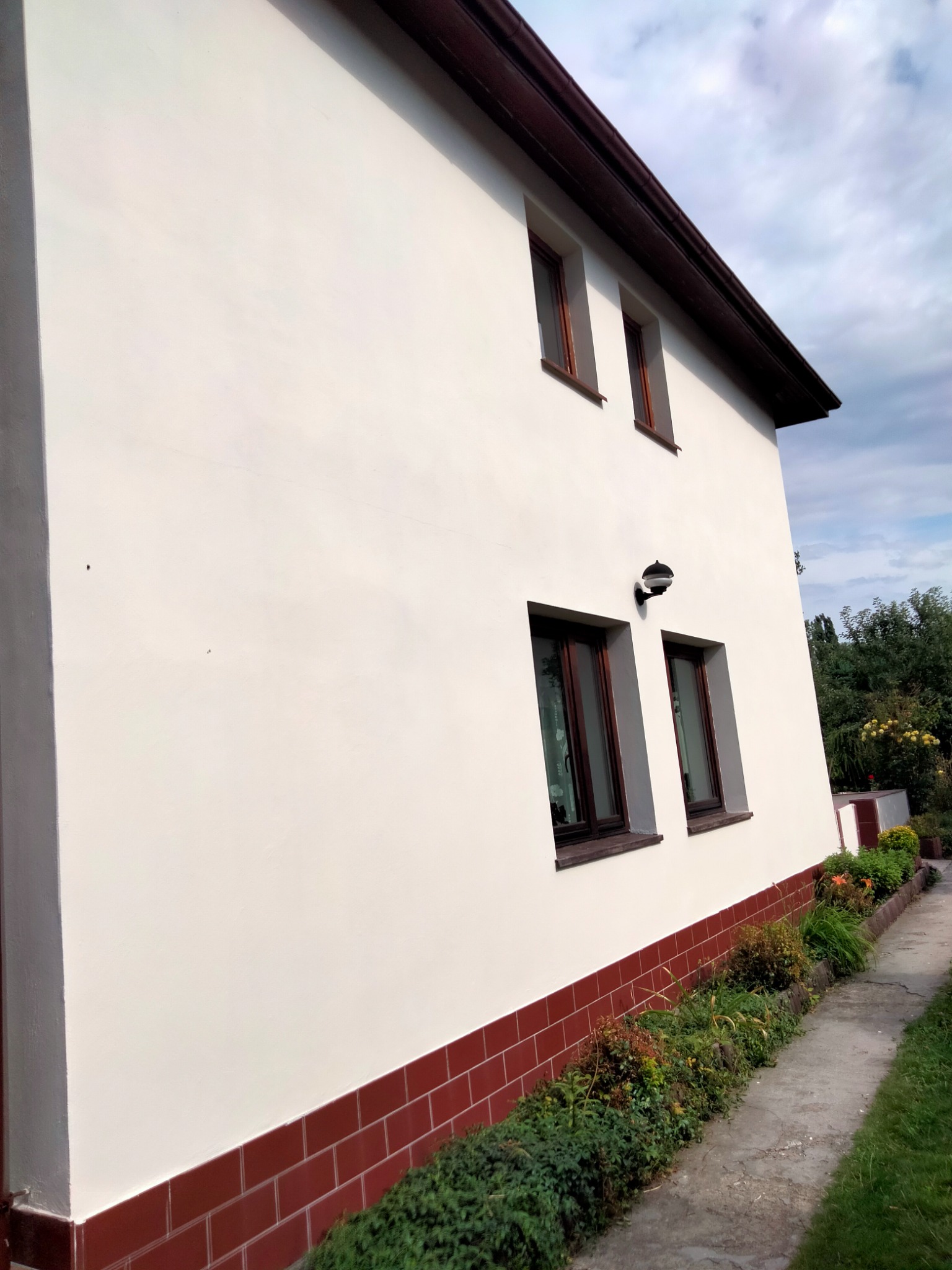 Boczna elewacja domu jednorodzinnego po remoncie, z nową, jasną farbą, brązowymi ramami okiennymi i czerwoną cegłą na fundamencie, widoczna z perspektywy chodnika.